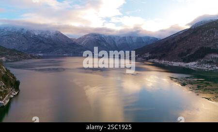 Vista aerea invernale di Gaupne, Norvegia, che mostra un fiordo che riflette la luce dorata, circondato da montagne innevate, un cielo nuvoloso e un tranquillo paesaggio rurale. Foto Stock