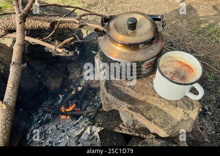 Tazza in smalto bianco riempita di bevanda calda accanto al bollitore nero rustico sulla fossa del fuoco in pietra. Nelle vicinanze, alcuni bastoncini sono disposti per il fuoco, il tutto sullo sfondo di un'area campeggio verde e boscosa. Foto Stock