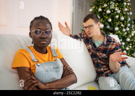 Giovane coppia multietnica che durante le festività festive ha litigato mentre si siede su un divano vicino all'albero di natale, incontrando difficoltà di relazione e problemi di comunicazione Foto Stock