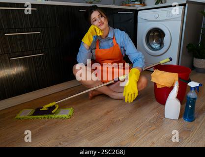 Donna stanca seduta sul pavimento, stanca di lavori domestici, prodotti per la pulizia e panni sparsi intorno. Foto Stock