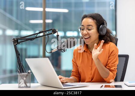 Blogger donna che registra podcast e corso di formazione online. Influencer che utilizza un microfono professionale e un laptop per lo streaming, seduto accanto alla finestra all'interno di un moderno studio. Foto Stock