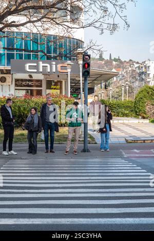 La gente sta aspettando il segnale stradale verde in una zebra pedonale. Un gruppo di persone di età diverse si trova all'attraversamento pedonale. Rosso l Foto Stock