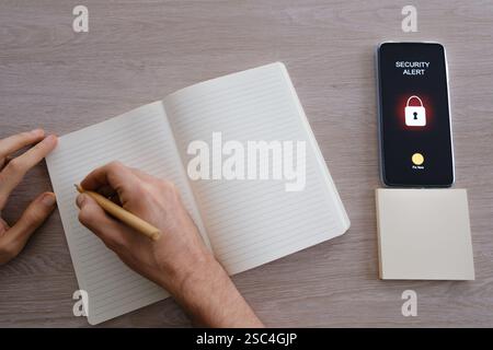 Persona che scrive nel notebook accanto allo smartphone e visualizza un avviso di sicurezza Foto Stock