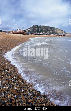 Le onde si infrangono dolcemente su una spiaggia di ciottoli, dove le barche da pesca riposano sullo sfondo vicino a una collina. L'atmosfera e' tranquilla, rivelando una zona costiera Foto Stock