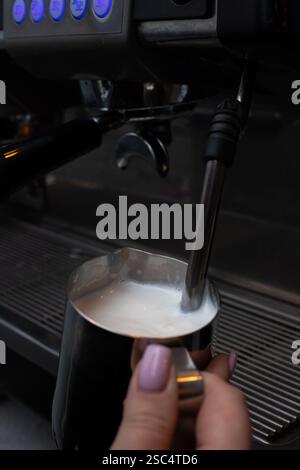 Primo piano delle mani del barista che montano il latte in una caraffa metallica utilizzando una macchina da caffè. Preparazione della schiuma di latte cremosa per cappuccino o latte. Foto Stock