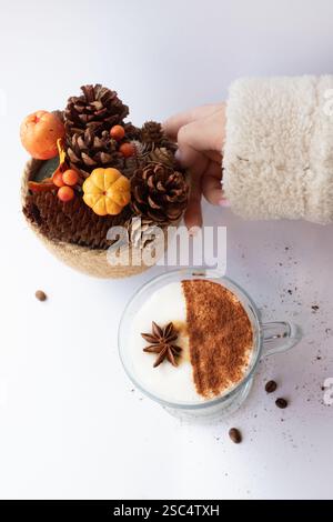 Accogliente ambiente autunnale con una mano in un morbido maglione che tocca una decorazione autunnale di coni di pino, frutti di bosco e una mini zucca accanto a un latte di spezie. Foto Stock