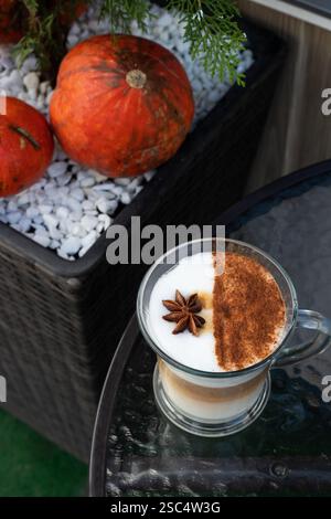 Latte autunnale con cannella e anice stellato su un tavolo di vetro. Intimo concetto autunnale con decorazioni di zucca sullo sfondo. Foto Stock
