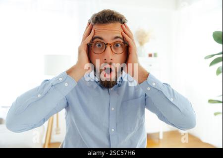 L'uomo con la barba sorpreso con gli occhiali in una camicia blu, tenendo la testa con entrambe le mani, esprimendo shock e stupore in un ufficio moderno e luminoso, guarda la fotocamera a bocca aperta Foto Stock