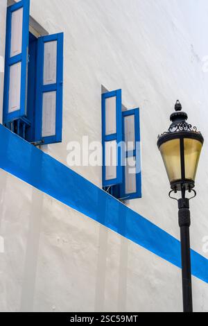 Le tende a finestra con cornice blu si aprono da un edificio dipinto di bianco su un'ampia banda blu su di una tradizionale lampada standard decorata. Foto Stock