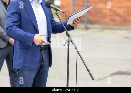 Uomo vestito con un blazer blu e jeans con i documenti, che tiene un discorso a un evento all'aperto mentre è in piedi vicino a un microfono Foto Stock