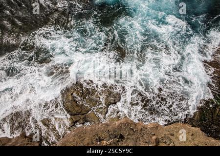 Vista aerea delle onde che si infrangono contro le rocce su un litorale. Pattern e texture dinamici nell'acqua e nelle rocce. Scena naturale potente e selvaggia. Foto Stock