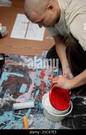 Immagine verticale del secchio in plastica aperto da un artista maschile con stampa a croce bianca su abiti con stencil di carta sul pavimento dell'officina macchiato Foto Stock