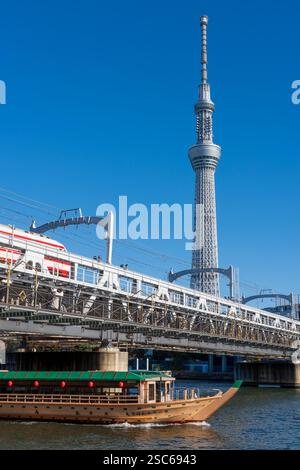 Passeggiata sul fiume Sumida, una barca sul fiume, un treno sul ponte ferroviario e il Tokyo Skytree sullo sfondo Foto Stock