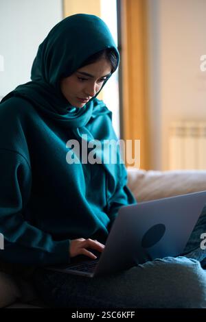 Giovane donna musulmana che indossa hijab che lavora da casa con un laptop Foto Stock