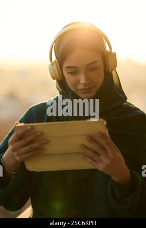Giovane donna musulmana che indossa le cuffie e usa un tablet al tramonto Foto Stock