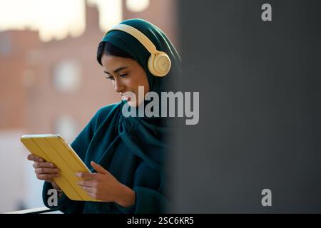 Giovane donna musulmana che ascolta musica e legge su un tablet all'aperto Foto Stock