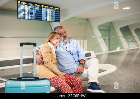 La coppia senior dorme pacificamente nell'abbraccio dell'altro su una panchina al terminal dell'aeroporto, in attesa della partenza del volo, incarnando l'essenc Foto Stock