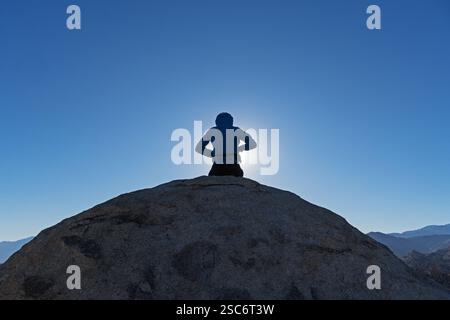 vista posteriore di una donna retroilluminata seduta sul masso in cima alla montagna con sfondo blu del cielo Foto Stock