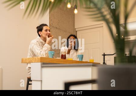 Due diverse donne sono sedute al bancone della cucina, mangiano toast e bevono caffè o succo di frutta, mentre si godono una mattinata rilassante nella loro moderna ed elegante h Foto Stock
