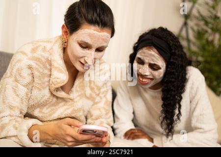 Due diverse amiche, vestite con comodi accappatoi e maschere facciali, sono sedute comodamente su un divano, rilassandosi e navigando sui social media sui loro Foto Stock