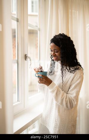 La giovane donna afroamericana, in comode poltrone, assapora una bevanda calda vicino a una finestra illuminata dal sole, creando un'atmosfera serena e accogliente nella sua casa Foto Stock