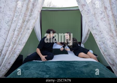 Coppia giovane felice che si gode una fuga romantica, seduti su un comodo letto all'interno di una tenda glamping, impegnati in una conversazione e condividendo un mese speciale Foto Stock