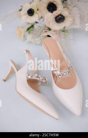 Le delicate scarpe da sposa bianche con punta a punta e un elegante design a fascia sono splendidamente esposte accanto a un bouquet di fiori su un morbido dorso blu Foto Stock