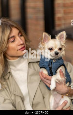 Una donna con lunghi capelli biondi culla delicatamente un piccolo cane bianco vestito con un gilet blu. Sono all'aperto, circondati da una vivace atmosfera cittadina. Foto Stock