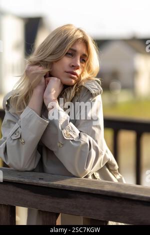 Una giovane donna dai capelli biondi si appoggia contro una ringhiera di legno, persa nel pensiero. Indossa un cappotto leggero e gode di un momento tranquillo in un vicino tranquillo Foto Stock