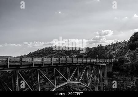 Un'impressionante fotografia in bianco e nero di un ponte in acciaio che si estende su un profondo canyon, con nuvole testurizzate sullo sfondo. L'immagine acquisisce Foto Stock