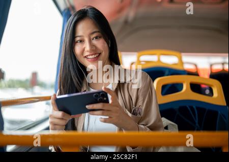 Una bella e allegra donna asiatica in abiti casual ama guardare un film o un video sul suo smartphone mentre siede davanti al finestrino su un autobus Foto Stock