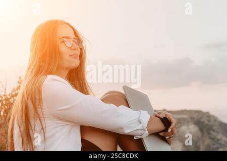 Donna laptop Sunset - donna sorridente con i capelli lunghi siede su una collina con un laptop e contempla il tramonto. Foto Stock
