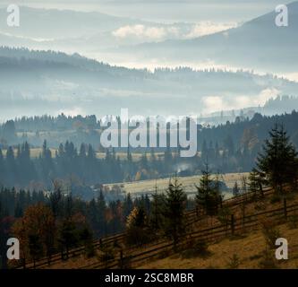 La luce del sole del mattino scorre attraverso la nebbia, illuminando un tranquillo villaggio di montagna annidato tra colline boscose. La luce crea raggi eterei che toccano delicatamente il fogliame autunnale e le case sparse sulle pendici. Foto Stock