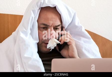 Uomo maturo malato seduto a letto con un portatile. Ha un fazzoletto nel naso Foto Stock