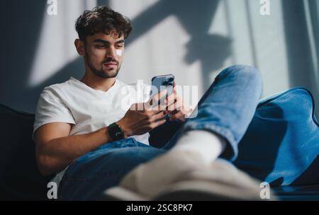 Un giovane uomo si rilassa a casa mentre utilizza uno smartphone in un moderno design Living Space Foto Stock