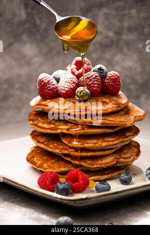 Frittelle con mirtilli e lamponi su un piatto con un cucchiaio con sopra il miele Foto Stock
