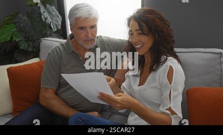 Coppia di mezza età seduta insieme in un accogliente soggiorno a leggere un documento con sorrisi condivisi e un'atmosfera calda che enfatizza l'amore e la festa Foto Stock