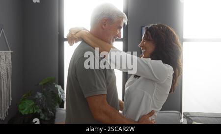 Coppia di mezza età che abbraccia nel soggiorno moderno, catturando amore e felicità all'interno con espressioni calde e gioiose circondate da interni accoglienti Foto Stock