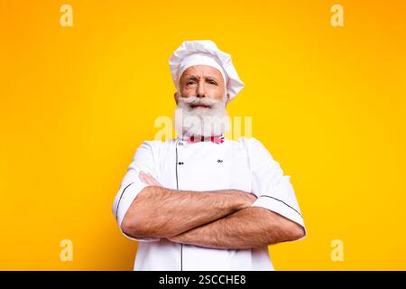 Ritratto di uno chef senior sicuro di sé che indossa una tradizionale uniforme con le braccia incrociate su un vivace sfondo giallo Foto Stock