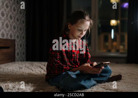 Ritratto di una ragazza concentrata seduta sul letto di casa, che utilizza lo smartphone per giocare o guardare video, circondato da un'atmosfera accogliente e buia Foto Stock