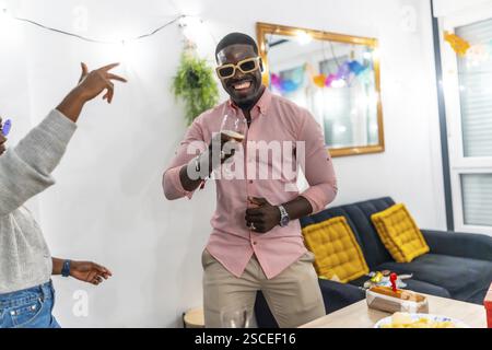 Uomo africano con t-shirt rosa e occhiali da sole sorridente bevendo birra e ballando godendosi una festa a casa con gli amici Foto Stock