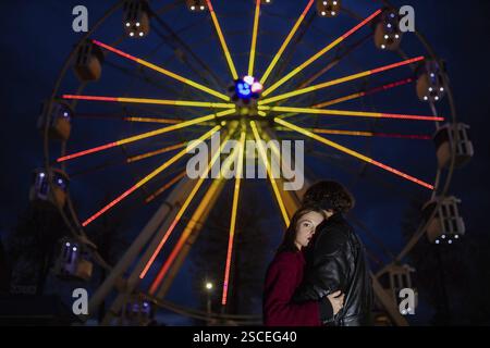 Una coppia si abbraccia sotto una ruota panoramica illuminata di notte Foto Stock