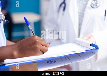 Infermiera afroamericana che tiene gli appunti e sbatte le informazioni essenziali sulla salute mentre il medico aspetta nelle vicinanze. Primo piano di lavoratrice sanitaria femminile nera che riempie i documenti medici nella stanza d'ospedale. Foto Stock