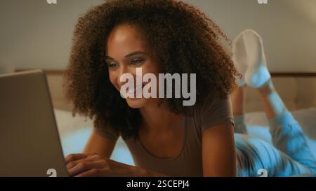 Donna afro-americana sorridente ragazza etnica che usa il laptop la sera sdraiata su un letto accogliente donna felice e allegra adolescente che digita sul computer online Foto Stock