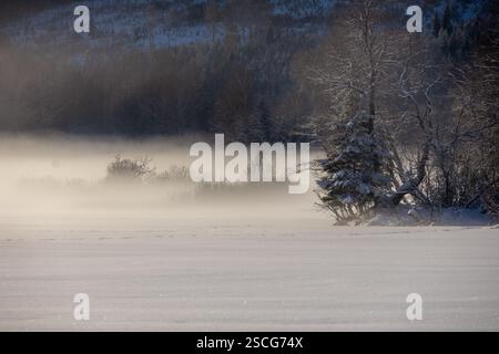 Un lago ghiacciato avvolto dalla nebbia, baciato dall'ultima luce del tramonto. Le montagne sono silenziose, mentre la bellezza dell'inverno si sviluppa in un abbraccio da sogno. Foto Stock