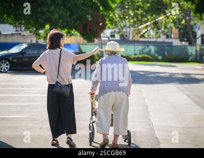 Donna anziana che cammina con un camminatore mobile nel parcheggio. Figlia adulta che adatta il cappello di madre anziana. California. Foto Stock