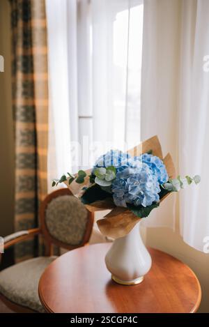 Un bouquet di ortensie blu avvolto in rustica carta kraft si trova graziosamente su un tavolo vicino alla finestra Foto Stock
