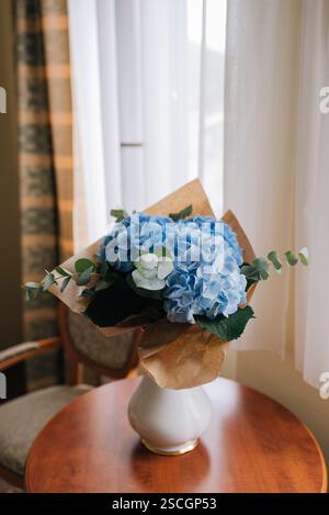 Un bouquet di ortensie blu avvolto in rustica carta kraft si trova graziosamente su un tavolo vicino alla finestra Foto Stock