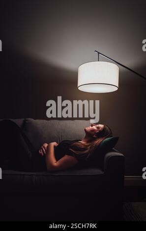Donna sdraiata sul divano in una stanza buia che guarda la luce della lampada. Foto Stock
