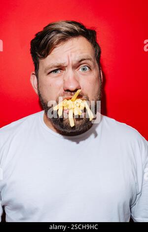 Un uomo barbuto con una t-shirt bianca ha una bocca piena di patatine fritte mentre fa un'espressione capricciosa. Il vivace sfondo rosso esalta il comico e. Foto Stock